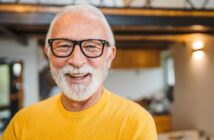 A smiling older man with white hair and a beard wears black-framed glasses and a bright yellow sweater. He stands in a warmly lit room with wooden accents and modern lighting in the background, creating a cozy atmosphere.