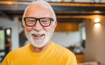 A smiling older man with white hair and a beard wears black-framed glasses and a bright yellow sweater. He stands in a warmly lit room with wooden accents and modern lighting in the background, creating a cozy atmosphere.