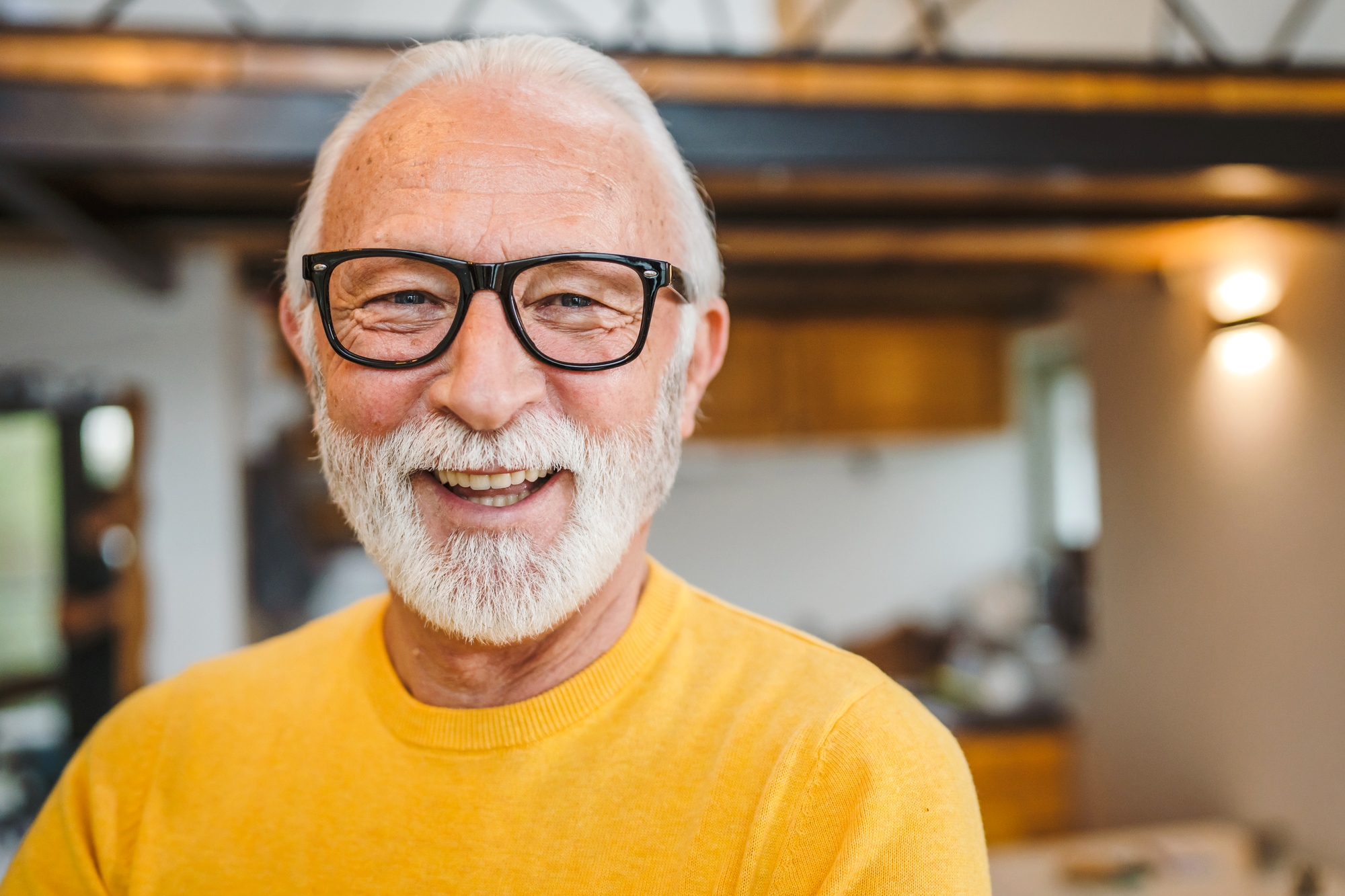 A smiling older man with white hair and a beard wears black-framed glasses and a bright yellow sweater. He stands in a warmly lit room with wooden accents and modern lighting in the background, creating a cozy atmosphere.