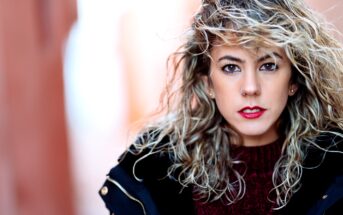 A woman with wavy blonde hair and red lipstick is looking directly at the camera. She is wearing a dark coat over a maroon sweater. The background is softly blurred, emphasizing her face and expression.