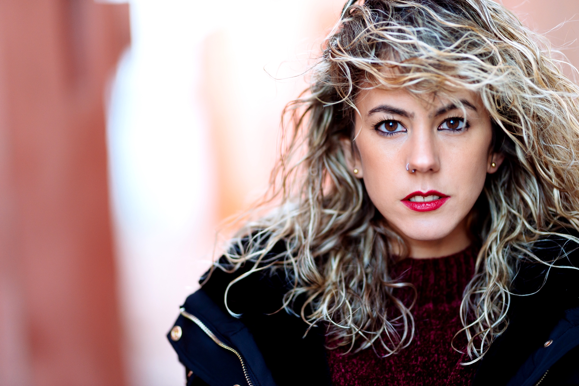 A woman with wavy blonde hair and red lipstick is looking directly at the camera. She is wearing a dark coat over a maroon sweater. The background is softly blurred, emphasizing her face and expression.