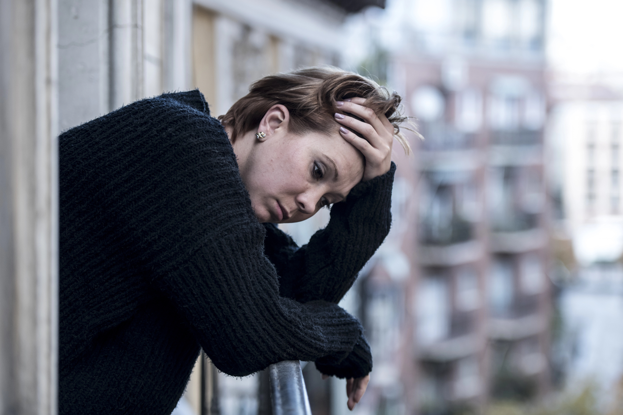 A person with short hair, wearing a dark sweater, leans on a balcony railing while holding their head with one hand. They appear contemplative or worried, with a blurred background of buildings and a cloudy sky.