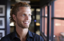 A smiling man with blue eyes and curly brown hair is looking towards a window in a brightly lit indoor setting. He is wearing a dark-colored shirt. The background is softly blurred, featuring framed pictures and a red wall.