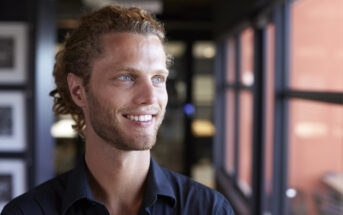 A smiling man with blue eyes and curly brown hair is looking towards a window in a brightly lit indoor setting. He is wearing a dark-colored shirt. The background is softly blurred, featuring framed pictures and a red wall.