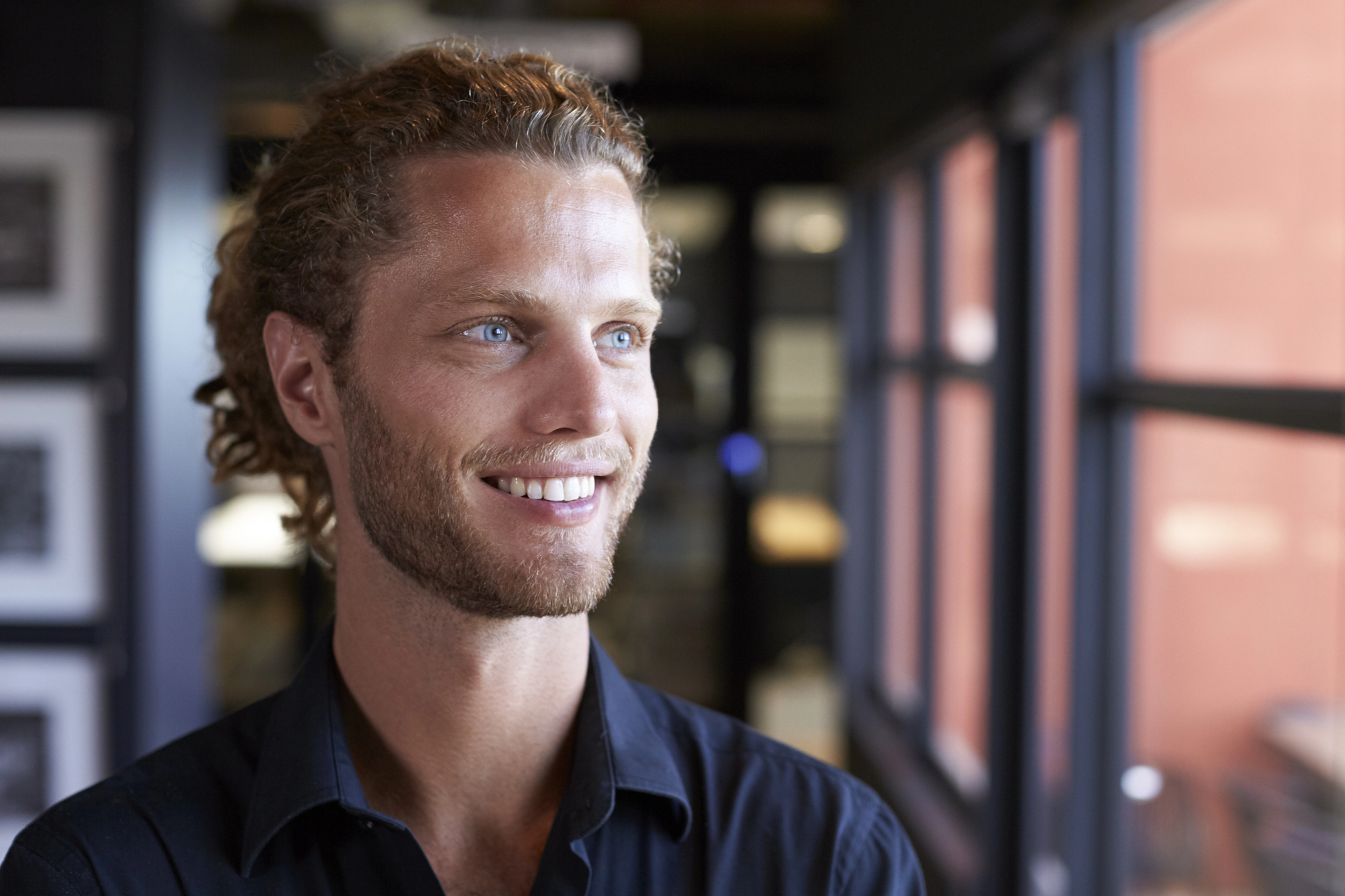 A smiling man with blue eyes and curly brown hair is looking towards a window in a brightly lit indoor setting. He is wearing a dark-colored shirt. The background is softly blurred, featuring framed pictures and a red wall.