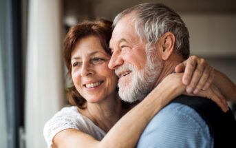 An older couple is embracing and smiling joyfully. The woman, with short brown hair, has her arms around the man's shoulders, while the man, with gray hair and a beard, leans into her, creating a tender and happy moment near a window with soft light.