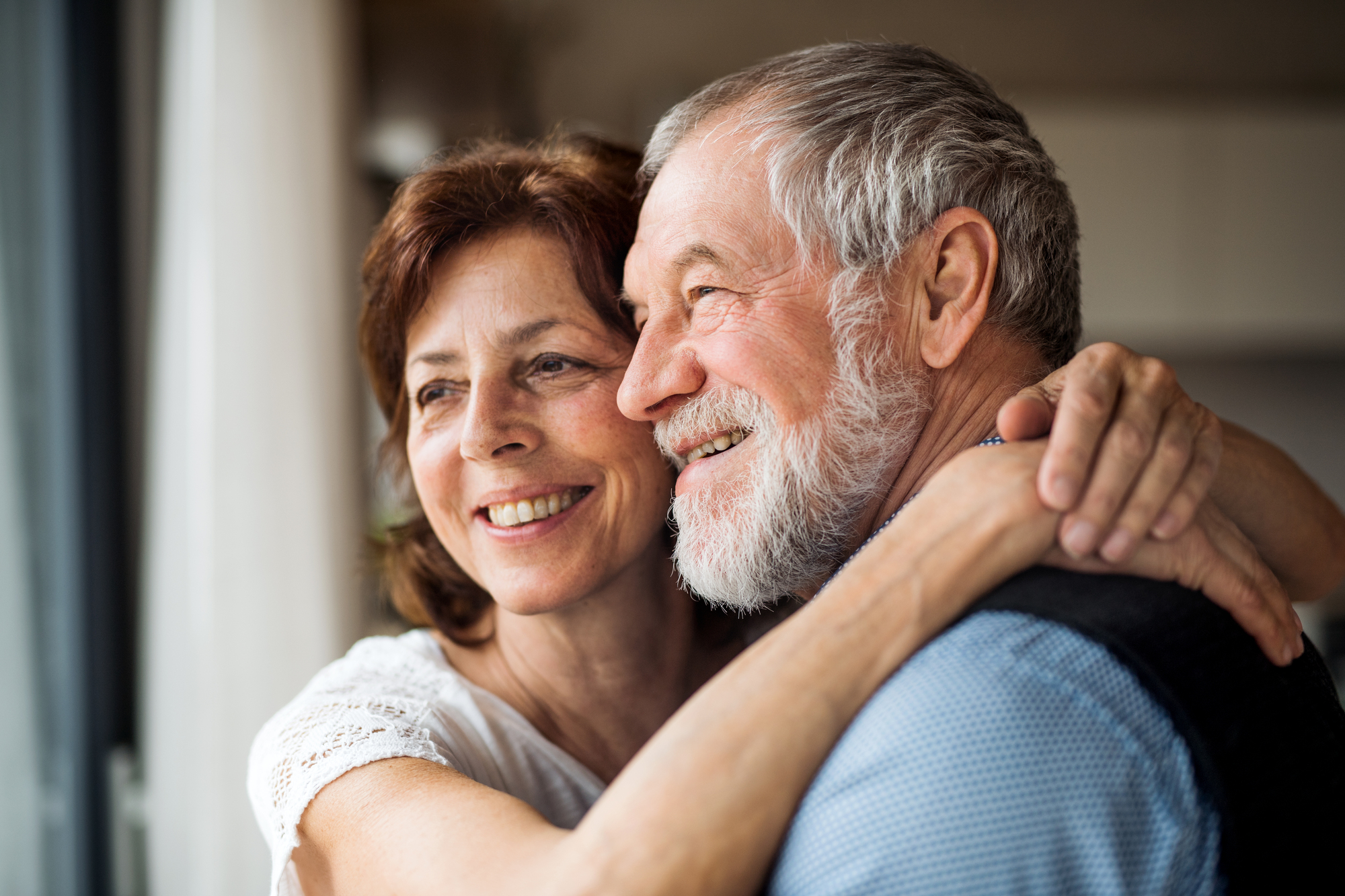 An older couple is embracing and smiling joyfully. The woman, with short brown hair, has her arms around the man's shoulders, while the man, with gray hair and a beard, leans into her, creating a tender and happy moment near a window with soft light.