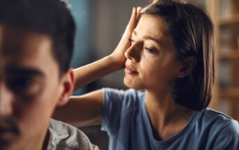 A woman with short dark hair and wearing a blue shirt rests her hand on her forehead while looking at a man in the foreground who is facing away from her. The scene suggests a moment of contemplation or concern.