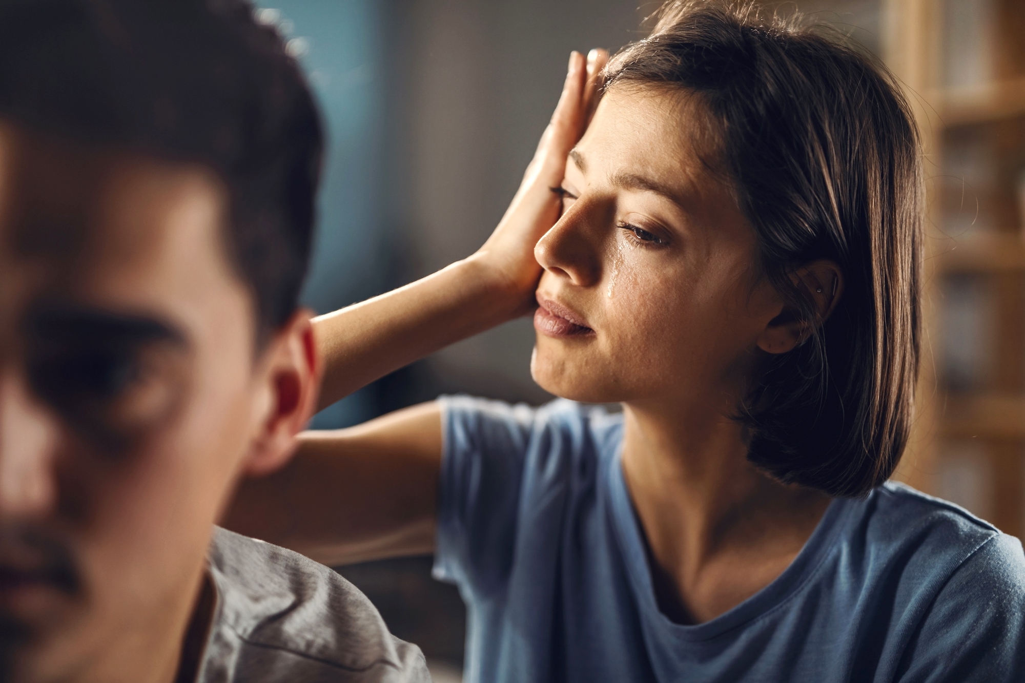 A woman with short dark hair and wearing a blue shirt rests her hand on her forehead while looking at a man in the foreground who is facing away from her. The scene suggests a moment of contemplation or concern.