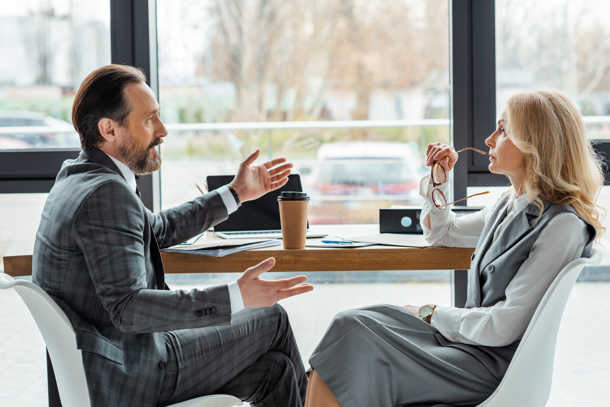 A man and woman in business attire sit across from each other at a table in an office. The man gestures with both hands while talking, and the woman listens while holding her glasses near her mouth. Laptops, documents, and a coffee cup are on the table.