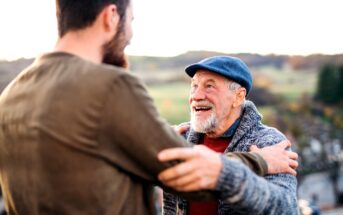 A joyous elderly man with a white beard, wearing a blue flat cap and gray cardigan, holds the arms of a younger man in a brown sweater. They are outdoors, surrounded by greenery and trees, sharing a heartfelt moment and smiling at each other.