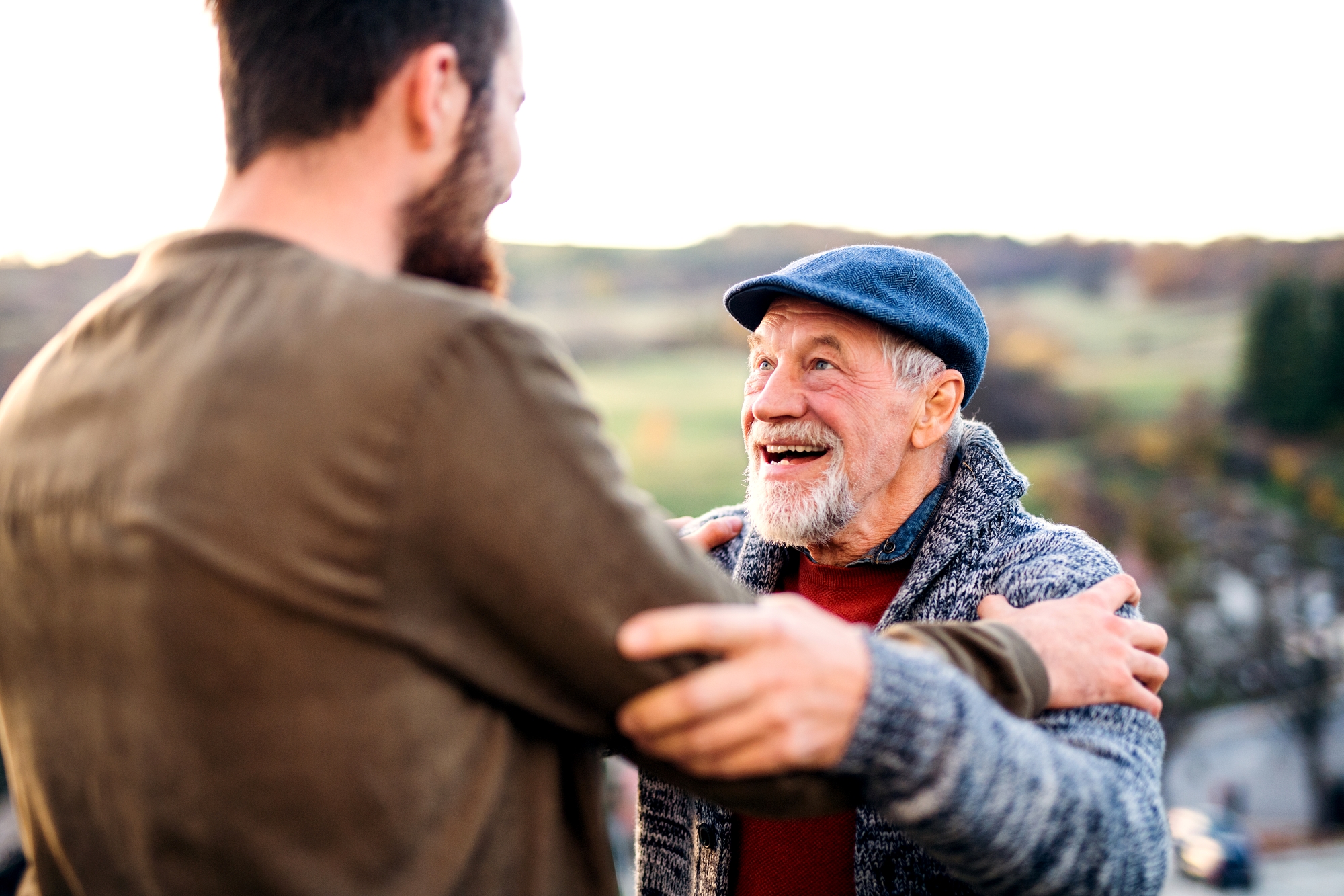 A joyous elderly man with a white beard, wearing a blue flat cap and gray cardigan, holds the arms of a younger man in a brown sweater. They are outdoors, surrounded by greenery and trees, sharing a heartfelt moment and smiling at each other.