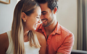 A smiling couple leans close to each other, their faces almost touching. The woman has long blonde hair, wears a white sleeveless top, and gold earrings. The man, with dark hair and a beard, is in a salmon-colored shirt. Both look very happy. The background is softly lit.