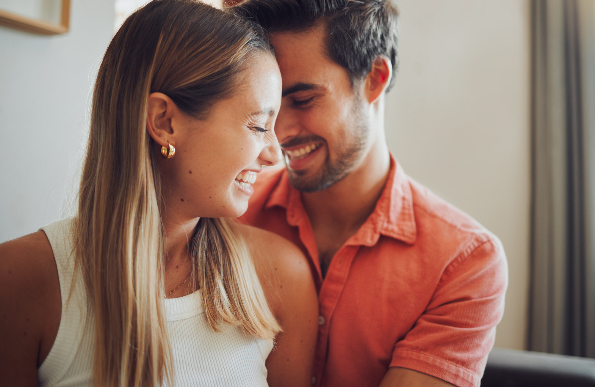 A smiling couple leans close to each other, their faces almost touching. The woman has long blonde hair, wears a white sleeveless top, and gold earrings. The man, with dark hair and a beard, is in a salmon-colored shirt. Both look very happy. The background is softly lit.