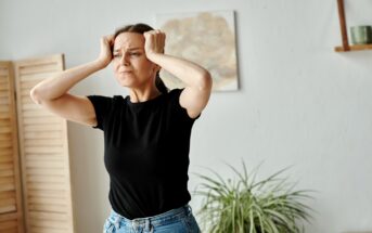 A woman in a black t-shirt and blue jeans stands indoors, holding her head with both hands, appearing frustrated or stressed. A wooden room divider, a wall painting, and a plant are visible in the background.