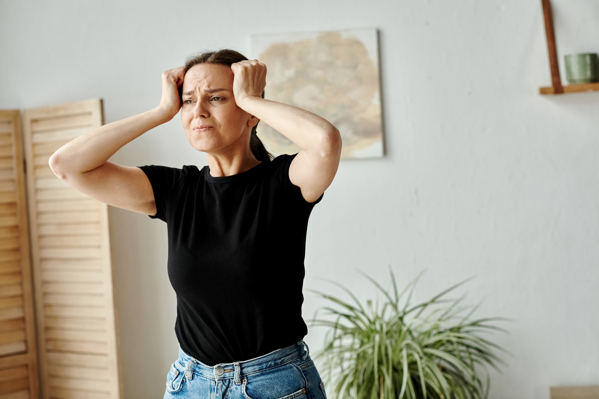 A woman in a black t-shirt and blue jeans stands indoors, holding her head with both hands, appearing frustrated or stressed. A wooden room divider, a wall painting, and a plant are visible in the background.