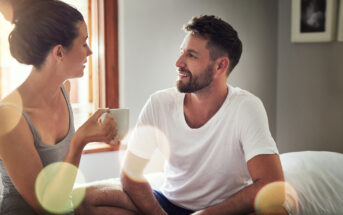 A man and woman are sitting indoors, talking and smiling at each other. The woman is holding a white mug, while the man is sitting on a bed. Soft, natural light is streaming in through a window, and there are lens flare effects in the image.