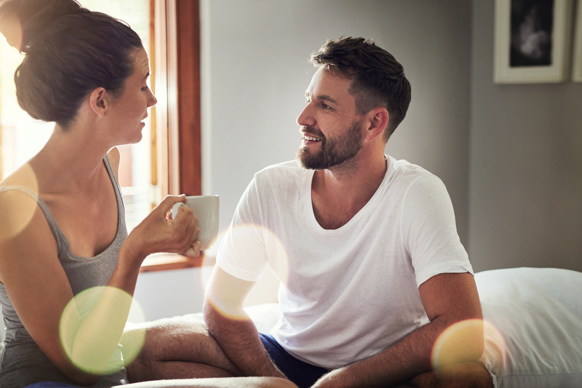 A man and woman are sitting indoors, talking and smiling at each other. The woman is holding a white mug, while the man is sitting on a bed. Soft, natural light is streaming in through a window, and there are lens flare effects in the image.