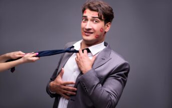 A man in a gray suit with lipstick marks on his face looks worried as he holds his tie, which is being pulled by a woman's hand. The background is plain gray.