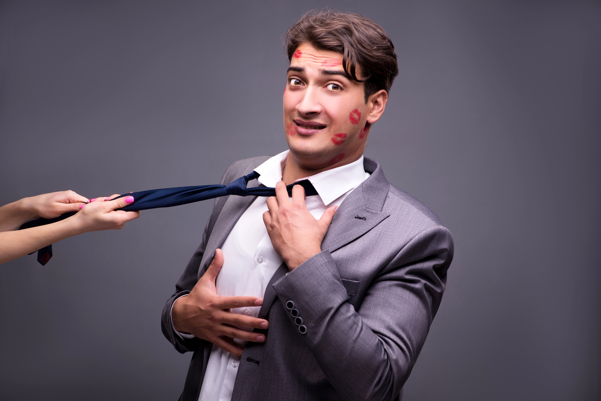 A man in a gray suit with lipstick marks on his face looks worried as he holds his tie, which is being pulled by a woman's hand. The background is plain gray.