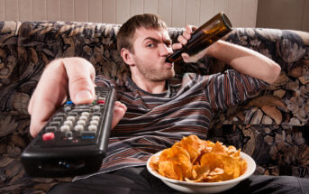 A man lounges on a patterned sofa, holding a remote control towards the camera in one hand and drinking from a beer bottle with the other. A bowl of potato chips rests on his lap. He wears a striped shirt and appears relaxed.