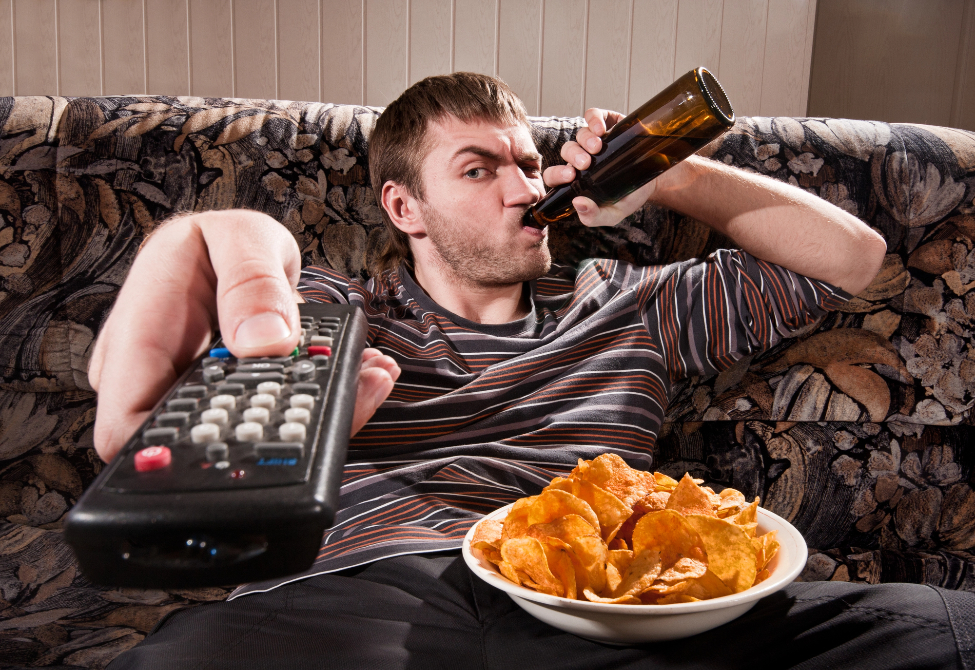 A man lounges on a patterned sofa, holding a remote control towards the camera in one hand and drinking from a beer bottle with the other. A bowl of potato chips rests on his lap. He wears a striped shirt and appears relaxed.