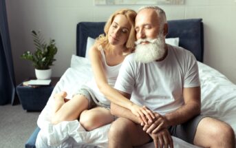 A woman with blonde hair and a man with a white beard are sitting on a bed in a cozy bedroom. Both are dressed in casual white and grey clothing. The woman sits behind the man, gently resting her head on his shoulder with her eyes closed. A plant is visible in the background.