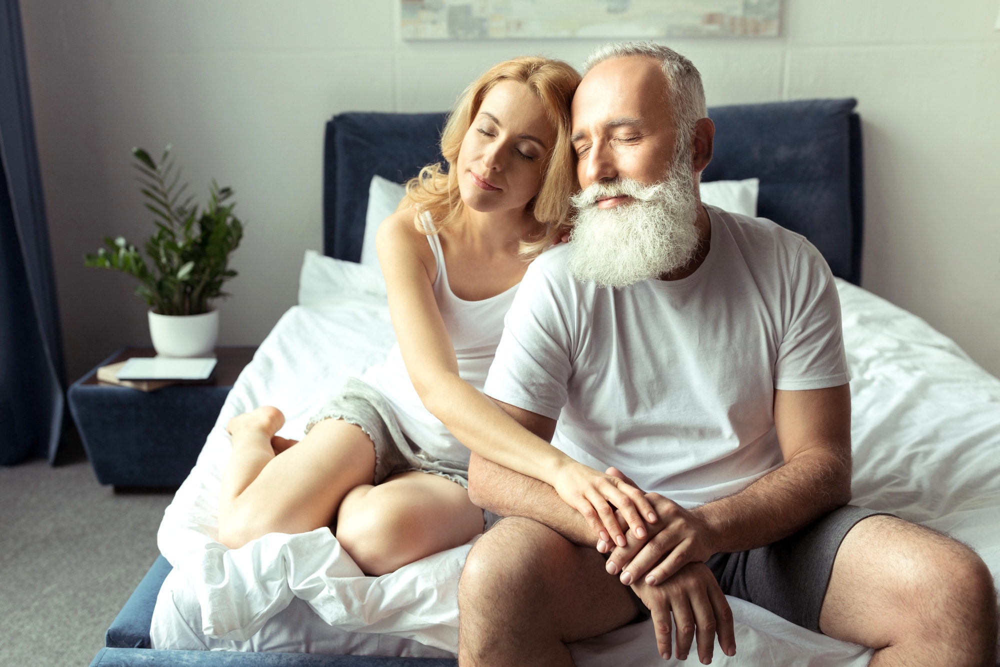 A woman with blonde hair and a man with a white beard are sitting on a bed in a cozy bedroom. Both are dressed in casual white and grey clothing. The woman sits behind the man, gently resting her head on his shoulder with her eyes closed. A plant is visible in the background.