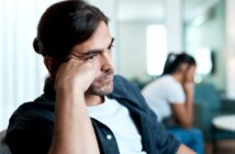 A man with long dark hair, beard, and mustache, wearing a white t-shirt and dark shirt, rests his head on his hand and looks thoughtful. A blurred figure of another person in the background also appears upset. The setting seems to be indoors.