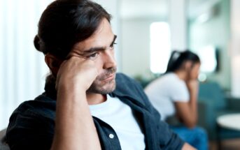 A man with long dark hair, beard, and mustache, wearing a white t-shirt and dark shirt, rests his head on his hand and looks thoughtful. A blurred figure of another person in the background also appears upset. The setting seems to be indoors.