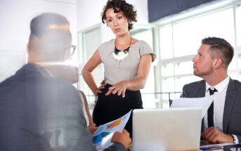 A woman stands at a table discussing something with a group of colleagues in a bright office. She is gesturing with her hands while the others, seated, listen and look at documents. A laptop sits open on the table in front of one of the seated men.