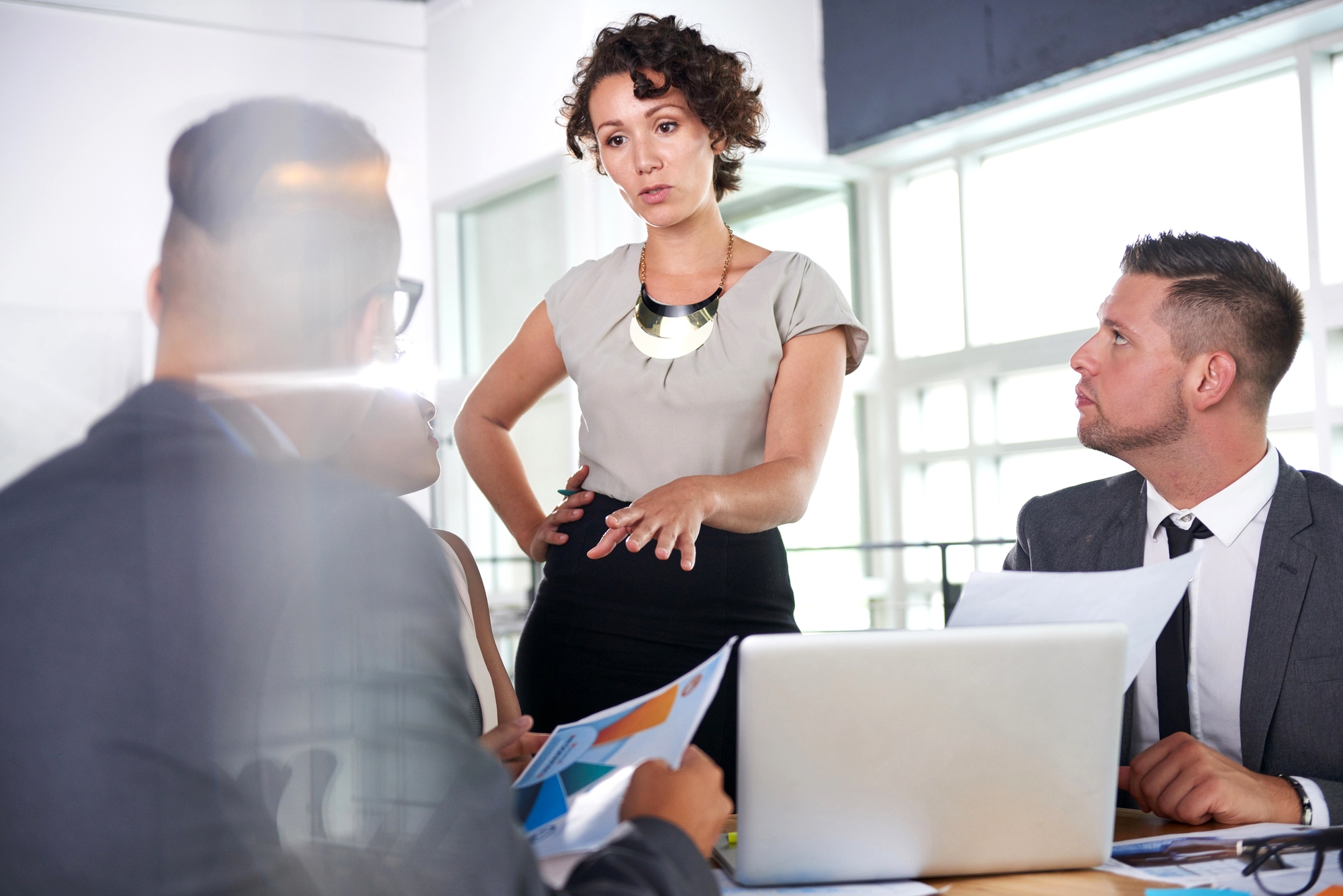 A woman stands at a table discussing something with a group of colleagues in a bright office. She is gesturing with her hands while the others, seated, listen and look at documents. A laptop sits open on the table in front of one of the seated men.