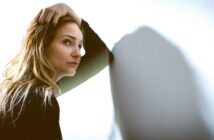 A woman with long, blonde hair leans against a white wall, her right hand running through her hair. She gazes pensively to her left, casting a soft shadow on the wall. She wears a black long-sleeve top, and the lighting is soft and natural.