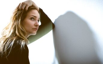 A woman with long, blonde hair leans against a white wall, her right hand running through her hair. She gazes pensively to her left, casting a soft shadow on the wall. She wears a black long-sleeve top, and the lighting is soft and natural.