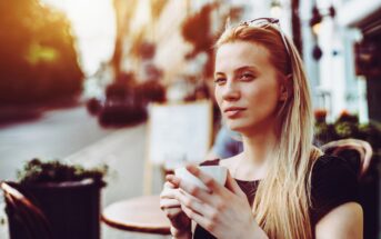 A woman with long blonde hair sits at an outdoor café, holding a white coffee cup. She wears sunglasses on her head and looks relaxed, with a soft smile. The background shows a blurred street scene with warm, golden sunlight filtering through.