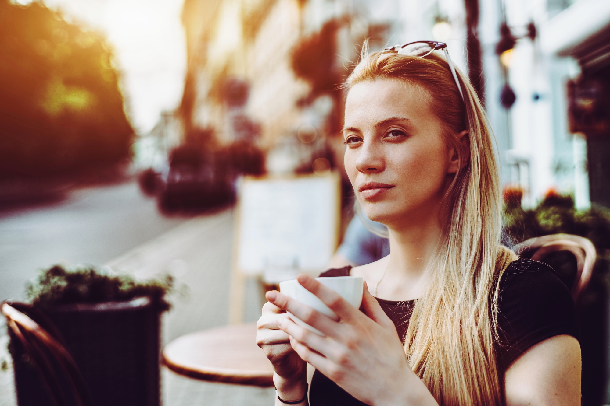 A woman with long blonde hair sits at an outdoor café, holding a white coffee cup. She wears sunglasses on her head and looks relaxed, with a soft smile. The background shows a blurred street scene with warm, golden sunlight filtering through.