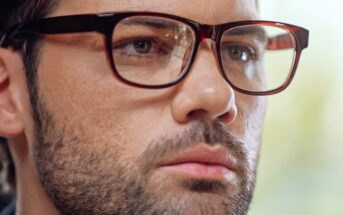 A close-up image of a person with short dark hair and a beard, wearing glasses with dark red frames. The individual has a serious expression, and the background is blurred.