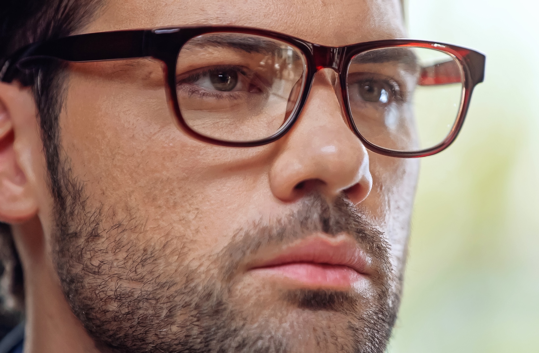 A close-up image of a person with short dark hair and a beard, wearing glasses with dark red frames. The individual has a serious expression, and the background is blurred.