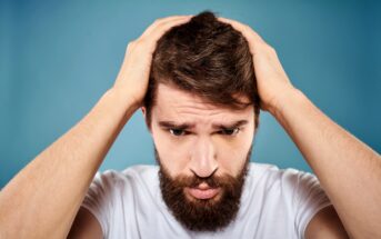 A bearded man wearing a white t-shirt stands against a blue background, holding his head with both hands and looking downward with a troubled expression.