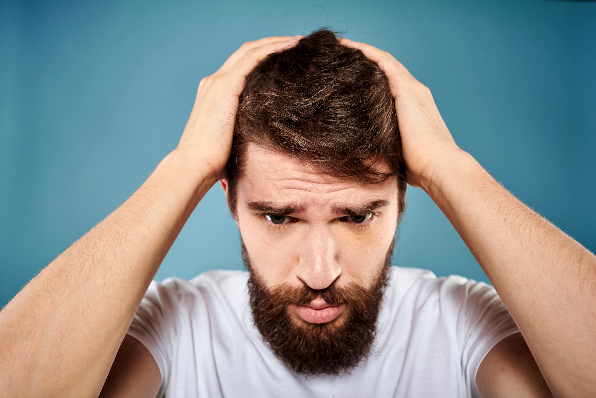 A bearded man wearing a white t-shirt stands against a blue background, holding his head with both hands and looking downward with a troubled expression.