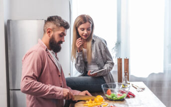 A bearded man in a pink shirt is chopping yellow bell peppers on a wooden cutting board in a modern kitchen. A woman with long blonde hair, in a gray shirt, is sitting on the counter beside him, holding a tablet and smiling. A bowl of fresh vegetables sits nearby.