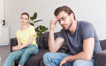 A man wearing a gray t-shirt and glasses sits on a couch with a distressed expression, resting his forehead on his fingers. A woman in a yellow shirt and light blue pants sits farther away, looking at him with concern. They are in a bright room with white walls.