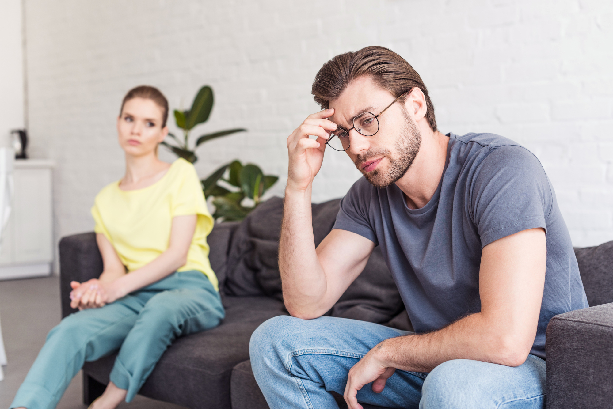 A man wearing a gray t-shirt and glasses sits on a couch with a distressed expression, resting his forehead on his fingers. A woman in a yellow shirt and light blue pants sits farther away, looking at him with concern. They are in a bright room with white walls.