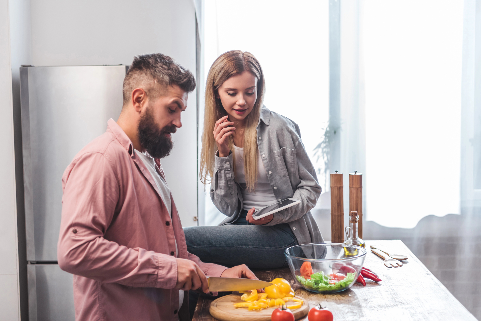 A bearded man in a pink shirt is chopping yellow bell peppers on a wooden cutting board in a modern kitchen. A woman with long blonde hair, in a gray shirt, is sitting on the counter beside him, holding a tablet and smiling. A bowl of fresh vegetables sits nearby.