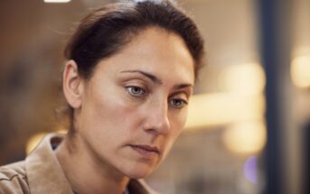 A woman with dark hair pulled back is looking down with a thoughtful expression. She is wearing a tan coat. The background is softly blurred with warm lighting.