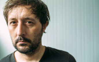 A man with short, dark, disheveled hair and a beard looks directly at the camera with a serious expression. He is wearing a plain black t-shirt, and the background is a light-colored, vertical-striped wall.