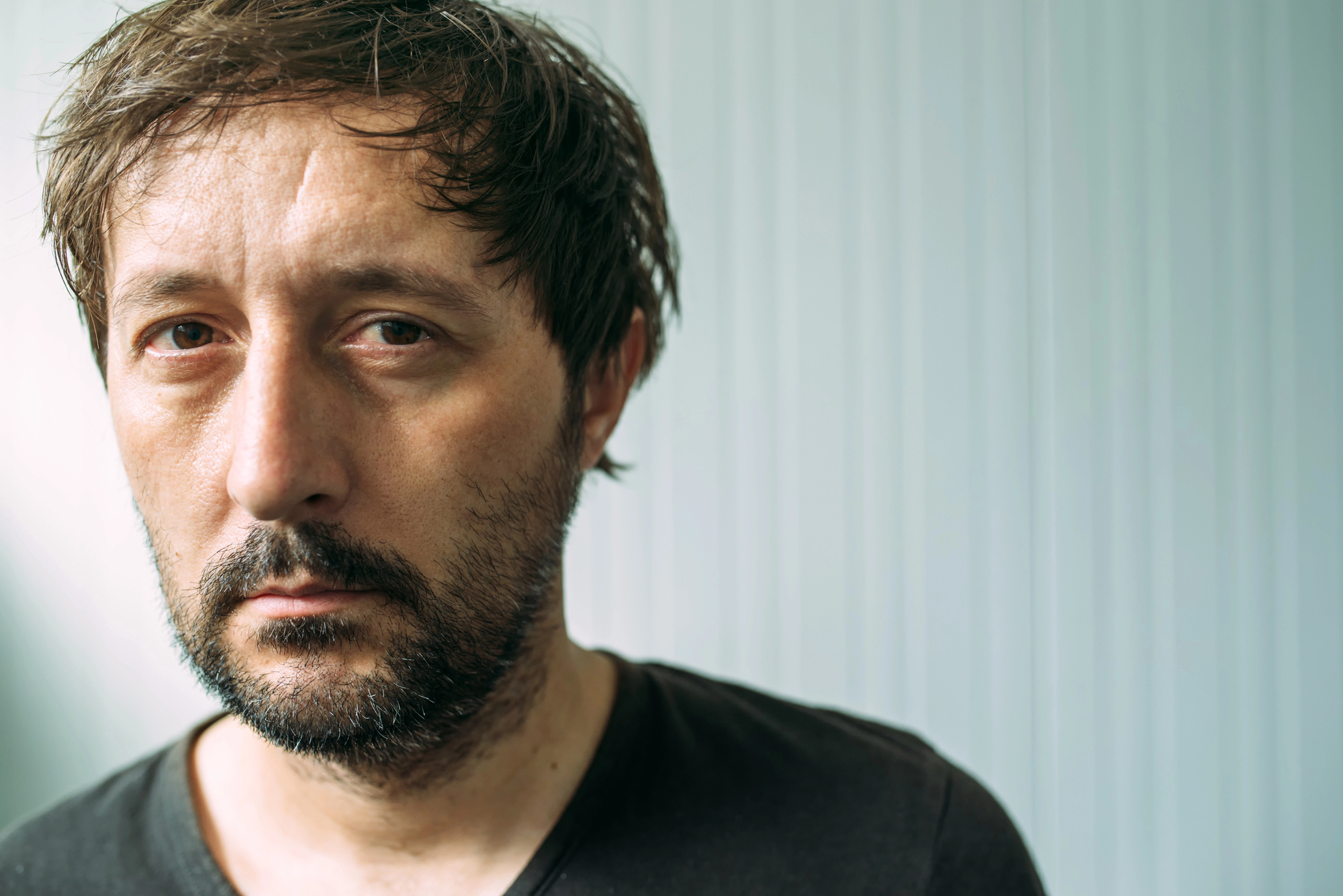 A man with short, dark, disheveled hair and a beard looks directly at the camera with a serious expression. He is wearing a plain black t-shirt, and the background is a light-colored, vertical-striped wall.