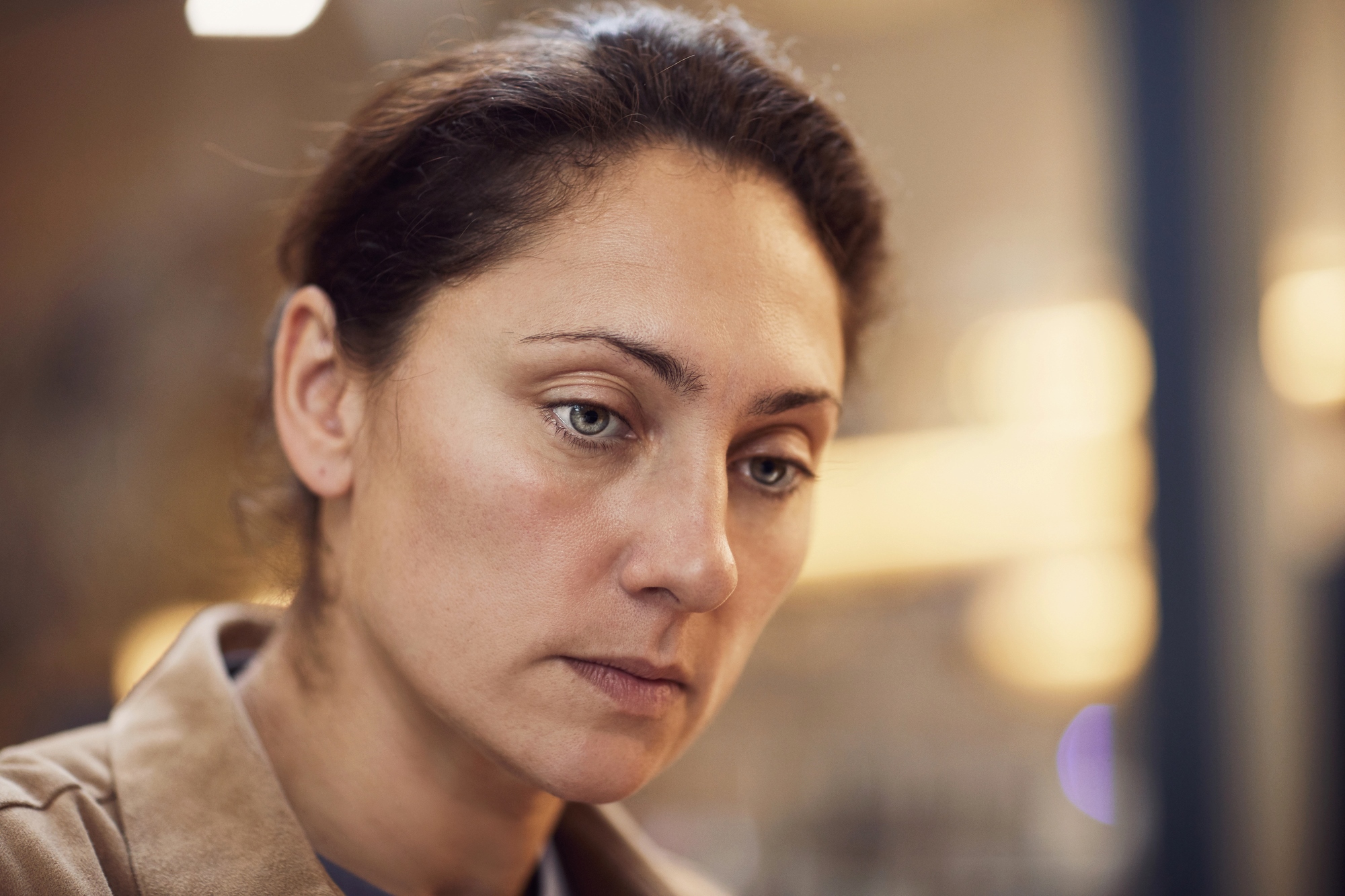 A woman with dark hair pulled back is looking down with a thoughtful expression. She is wearing a tan coat. The background is softly blurred with warm lighting.