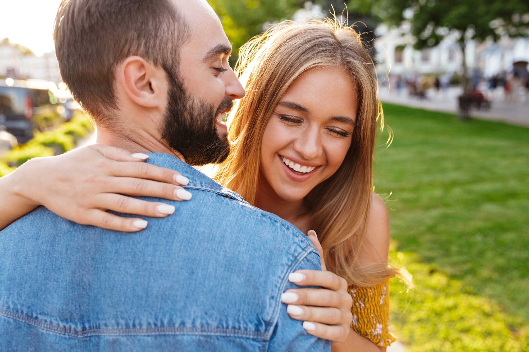 A young couple stands close together outdoors, smiling warmly at each other. The woman, with long blonde hair, embraces the man from behind, who has a beard and wears a denim jacket. The background features a grassy park and blurred buildings on a sunny day.