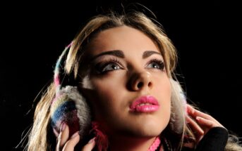 A woman with bright pink lipstick looks upward while wearing colorful, fluffy earmuffs. The background is black, highlighting her face and the earmuffs. Her expression is thoughtful or contemplative.